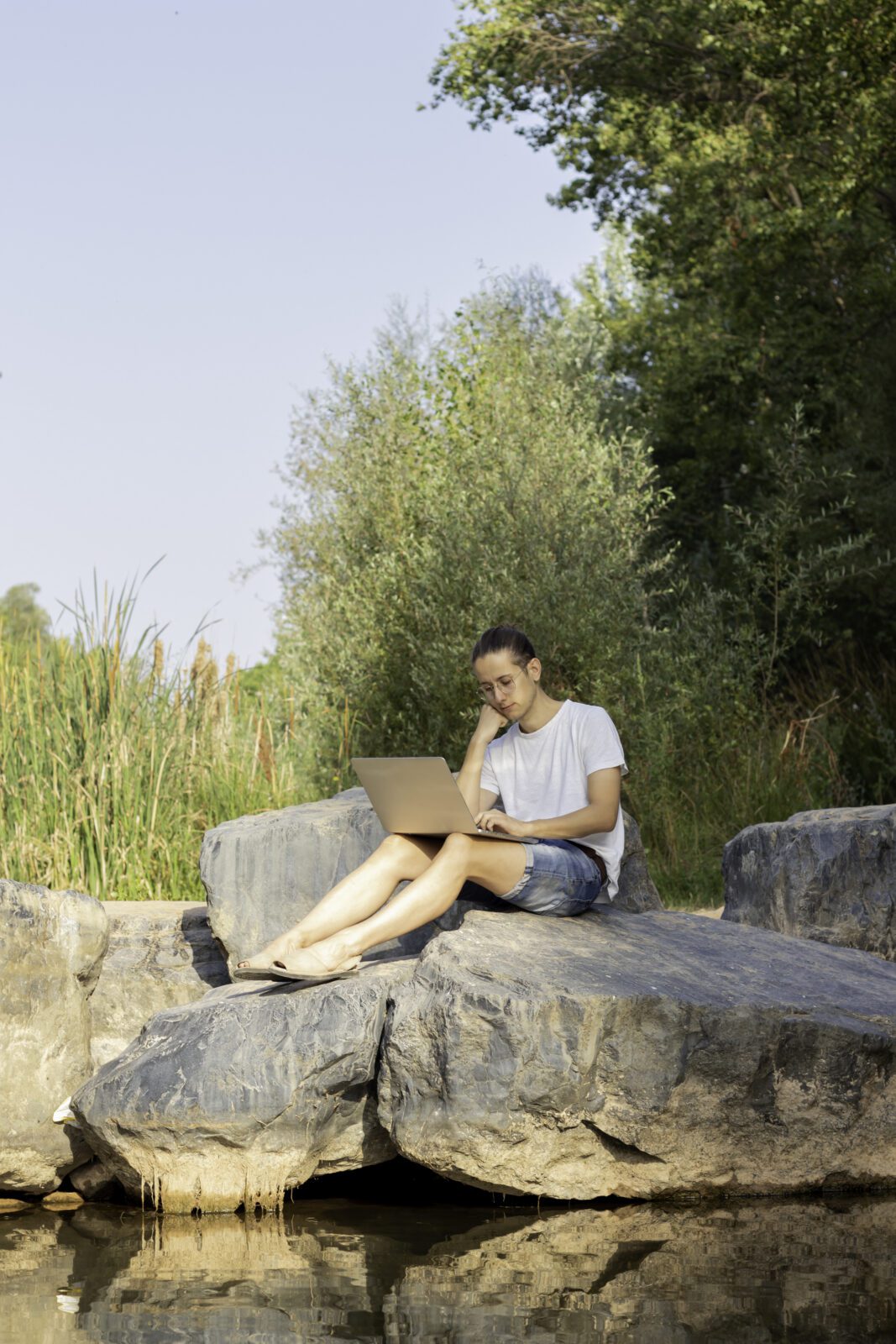 Vertical immagen of a young entrepreneur man sitting on the riverbank with a computer in his spare time green fern plant in close up photography